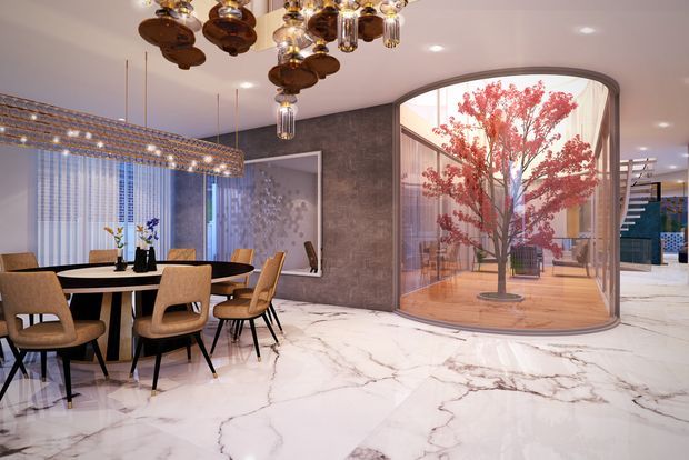 A luxurious contemporary dining and living area with a round dining table and upholstered chairs, a sculptural crystal and bronze pendant installation, polished white marble-effect flooring, dark textured wall finishes, and a striking curved glass enclosure containing an ornamental red-leaf tree. A stairway and additional interior spaces are visible beyond the glass.