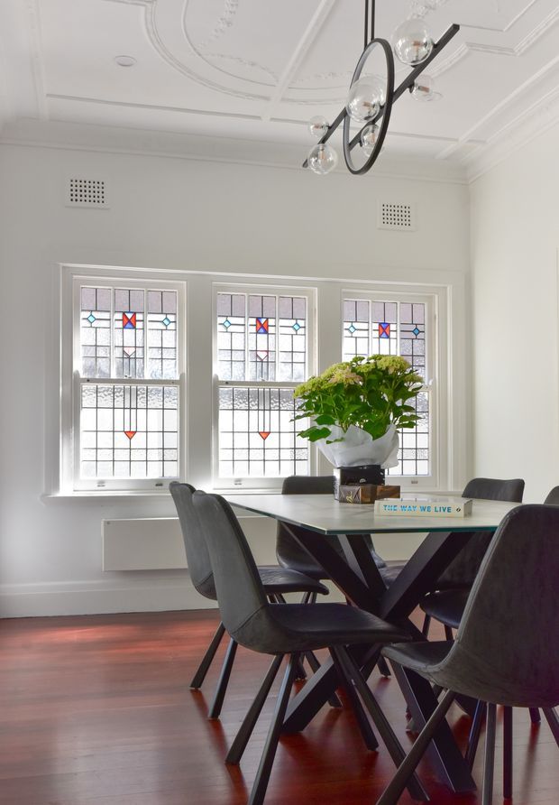 A bright dining room with a contemporary dining table and dark upholstered chairs, set against three decorative stained-glass windows. The room features polished dark timber flooring, white walls, ornate pressed ceiling detailing, decorative wall vents, and a sculptural black linear pendant light with globe bulbs. A large potted plant forms the centrepiece on the table.
