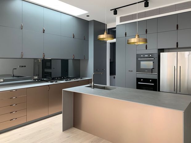 A contemporary kitchen with a large central island, extensive matte grey cabinetry, integrated black appliances, a stainless-steel refrigerator, pale timber flooring, and two brass pendant lights. The island includes a light stone-look benchtop and undermount sink, while linear ceiling vents and track lighting contribute to the streamlined design.