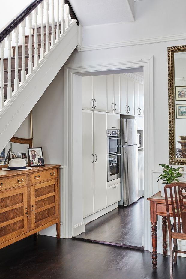 A refined traditional interior showing a dark timber-floored dining or living area beneath a staircase, opening directly into a compact white kitchen. The kitchen has full-height paneled cabinetry, integrated stainless-steel appliances and curved metal handles. White painted wall panelling, decorative mouldings, a turned stair balustrade, an ornate framed mirror, and a timber sideboard contribute to the classic character.
