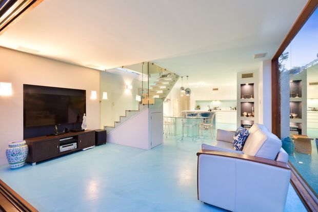 Open-plan contemporary living area with a large television and low dark entertainment unit, pale polished flooring, a light upholstered sofa, and a compact kitchen with island and transparent bar stools beyond. A glass balustrade staircase leads upward, while full-height glazing and timber-framed openings line the right side. Integrated display shelving and warm wall lighting add contrast to the bright, airy interior.