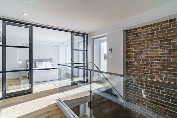 A bright contemporary interior landing overlooking a stairwell, with pale timber flooring, a transparent glass-and-dark-metal balustrade, black-framed internal glazing, and an exposed red-brown brick feature wall. A bedroom is clearly visible through the glazed partition.