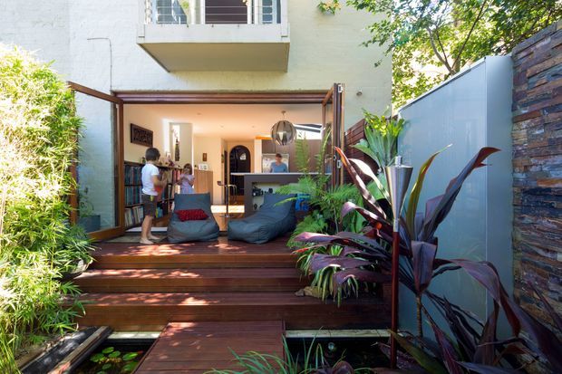 A lush, contemporary outdoor entertaining area viewed from a landscaped garden. A timber deck with broad steps leads into an open-plan interior through fully opened glazed doors, with beanbag seating and a small bar or kitchen area visible inside. Tropical planting, bamboo screening, a rendered multi-storey wall, metal privacy panel and stacked-stone feature wall frame the space. A narrow pond or water feature appears beneath the deck walkway.
