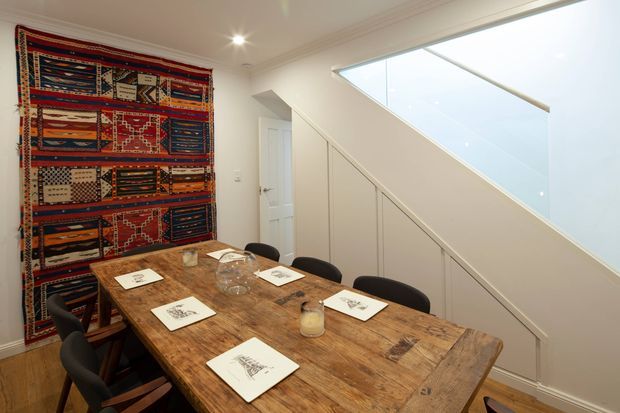 A bright, compact dining room with a long rustic timber dining table surrounded by dark upholstered chairs. A richly patterned textile wall hanging creates a bold feature wall, while a large triangular glazed stairwell opening and white built-in under-stair cabinetry add architectural interest. The room has white walls, ceiling, trim, and timber flooring, with recessed and spot lighting.