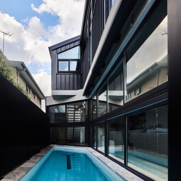 A narrow urban courtyard between a contemporary multi-storey residence and boundary fencing, centred on a long rectangular swimming pool. The home features extensive floor-to-ceiling glazing, dark metal-framed windows and doors, black vertical cladding, projecting upper volumes, and strong linear forms. Interior living areas are visible through the glazing, while the pool and paved edge dominate the foreground.