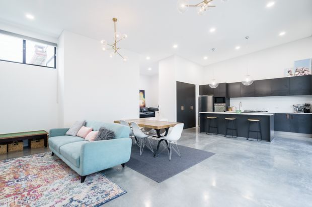 Bright open-plan interior combining a living room, dining area and contemporary kitchen. The space has polished pale concrete flooring, white walls and ceiling, dark kitchen cabinetry, a central island with stools, a timber dining table with white chairs, and a pale blue sectional sofa over layered area rugs. Recessed lighting and sculptural pendant fixtures contribute to a clean, spacious aesthetic.
