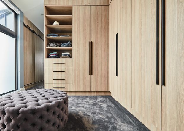 A contemporary walk-in wardrobe with extensive floor-to-ceiling light timber cabinetry, open shelving, drawers, and tall black handles. A large tufted upholstered ottoman sits in the foreground on a dark patterned carpet, beside full-height glazing with dark frames. The space is clean, highly organized, and softly lit with a refined, modern feel.