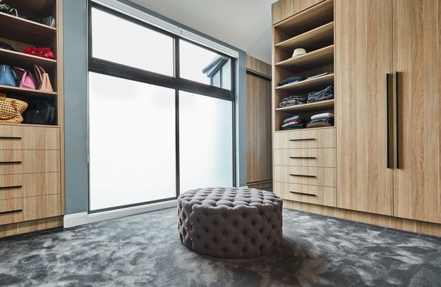 A spacious walk-in wardrobe or dressing room with floor-to-ceiling timber cabinetry, open shelving, drawers, hanging storage, and a central round tufted ottoman. Large black-framed glazing provides abundant natural light, while a patterned grey carpet covers the floor. The cabinetry has a warm, contemporary timber finish with black handles and trim.