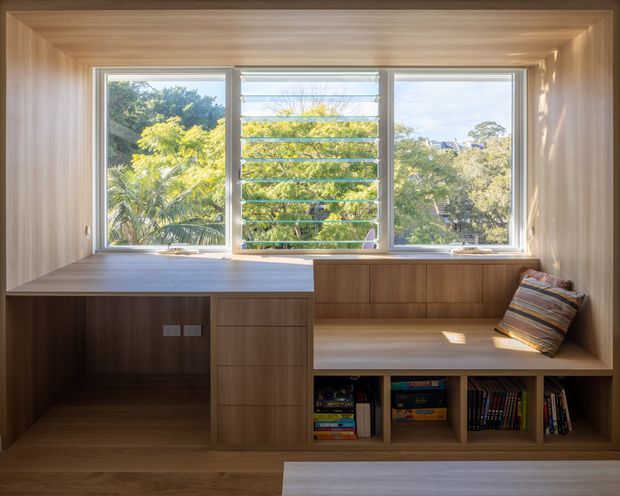 A compact built-in timber window nook combining a desk workspace on the left with a cushioned bench seat and open book shelving below. The full-height timber-lined reveal frames three windows, including a central louvred window, with abundant natural light and greenery visible outside. Integrated cabinetry and warm wood surfaces create a refined, space-efficient study or reading area.
