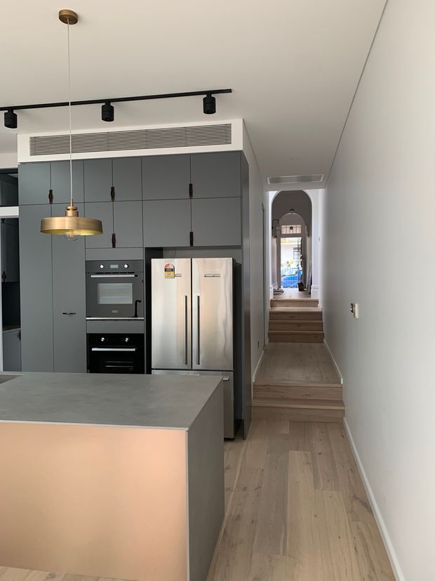 A contemporary kitchen viewed from the main living area, featuring a large grey island with a stone-look benchtop, full-height dark grey cabinetry, integrated black ovens, and a stainless-steel French-door refrigerator. A brass pendant light hangs above the island, while black track lighting and a linear ceiling vent add a modern industrial touch. Pale timber flooring continues through a narrow stepped hallway framed by white arched openings.