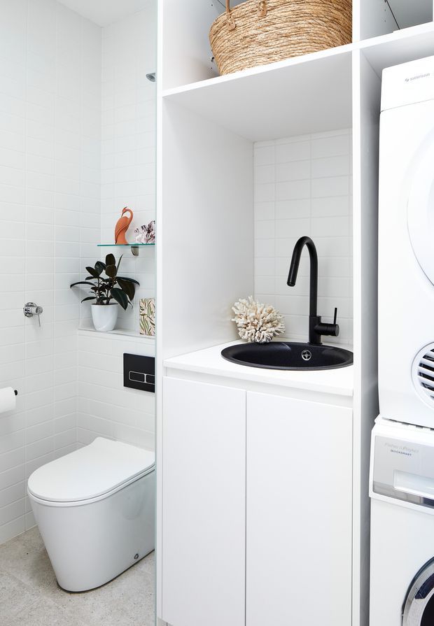 Bright, compact white utility bathroom/laundry with a wall-hung toilet, white tiled walls, a floating white vanity with black round basin and black tapware, open shelving with a woven basket, and a stacked white washing machine and dryer. Small decorative plants and coral add subtle natural accents.