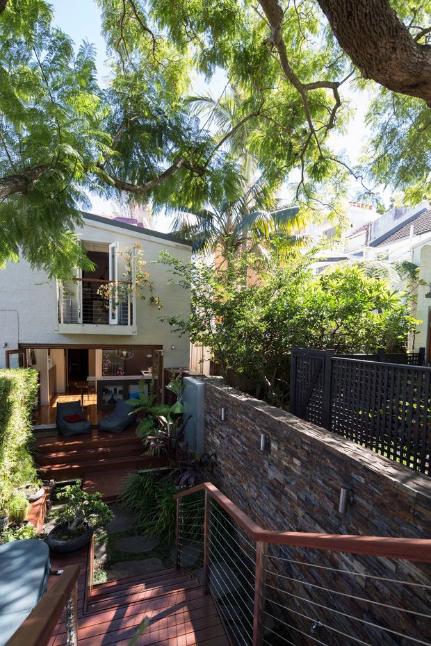 A bright, leafy courtyard and garden walkway beside a multi-level residential building. Timber decking and stairs descend through lush planting, with a dark wire balustrade and a textured stacked-stone retaining wall. The white rendered building has large glazed openings, an upper balcony with white-framed doors, and a tiled roof visible beyond the garden. The scene feels private, tropical and tranquil.