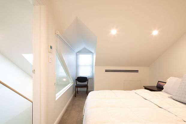 Bright compact bedroom with a low sloped ceiling, white bedding, neutral carpet, recessed downlights, and a narrow window seat or study nook at the far end. A large interior window or mirror-like glazed opening reflects the room, while a stair or landing is visible through the opening on the left.