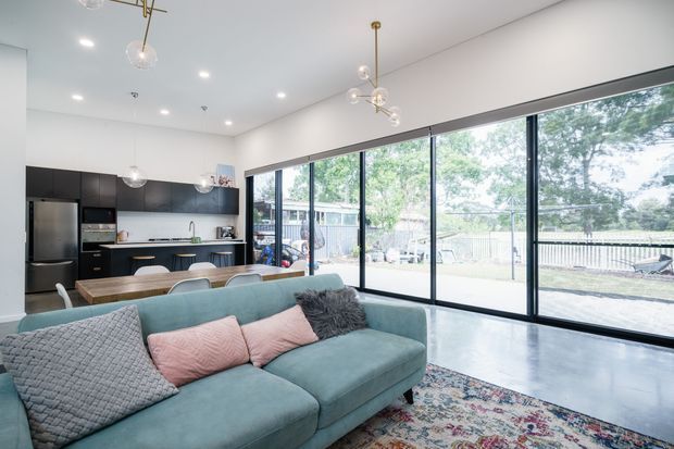 Bright open-plan living room with a contemporary kitchen at the rear, a large teal sectional sofa in the foreground, and extensive floor-to-ceiling sliding glass doors opening to an outdoor deck or yard. The room has polished concrete flooring, white walls and ceiling, dark kitchen cabinetry, timber island or dining surfaces, and decorative globe pendant lights.