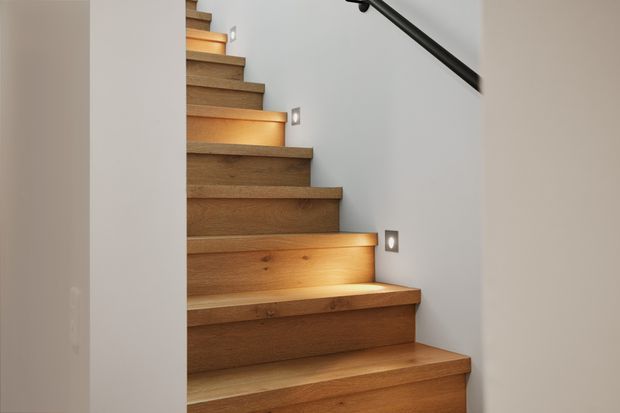 Close-up view of a modern timber staircase ascending between smooth white walls. The stairs have broad solid-wood treads and risers with integrated warm step lighting, while a simple black handrail runs along the upper right wall.