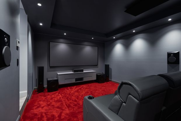 A dark, dedicated home theatre with a large wall-mounted projection screen, surround-sound speakers, recessed ceiling spotlights, a low media console, and black leather reclining seating. The room has dark grey walls and ceiling contrasted by a vivid red carpet, creating a dramatic cinematic atmosphere.