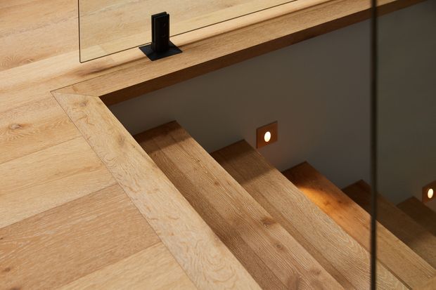 Close-up view looking down a timber staircase with broad natural wood treads and matching timber flooring. A frameless glass balustrade with dark metal hardware borders the stair opening, while small recessed wall lights illuminate the steps. The composition highlights warm wood grain, clean detailing, and a contemporary stair design.