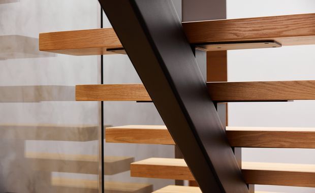 Close-up architectural detail of a modern stairway, showing thick warm timber treads and horizontal timber elements supported by a bold dark metal or painted structural beam. Clear glass balustrade and a light neutral wall create layered reflections and a clean, contemporary composition.