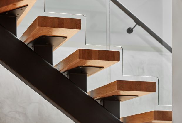 Close-up architectural view of a contemporary staircase with floating timber treads supported by dark metal or steel brackets and a substantial dark stair stringer. Thin clear or pale glass balustrade panels with dark metal fittings run alongside the stairs. The background includes a light grey concrete or plaster wall and a black track-style ceiling light, creating a refined, minimalist material palette.