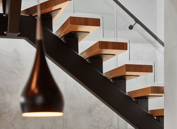 Close-up architectural view of a contemporary open staircase. Thick warm timber stair treads are supported by a dark steel structural stringer, with slim metal handrails and a sculptural dark pendant light in the foreground. The stair is set against a pale textured wall and appears to have a high ceiling or open vertical volume.