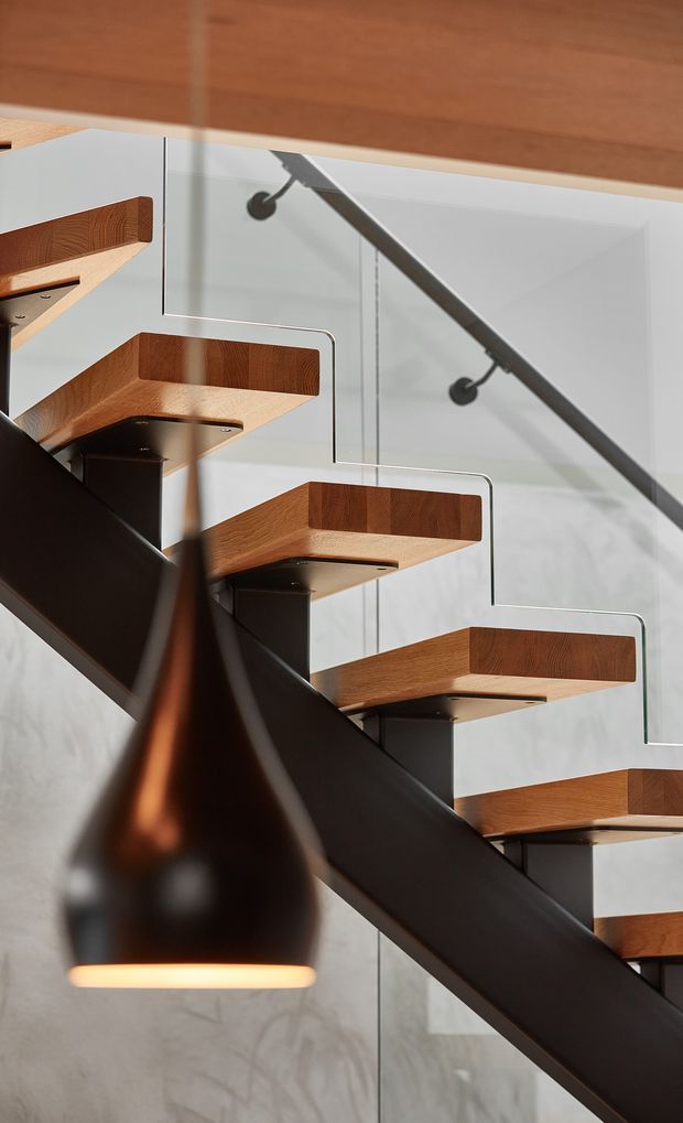 Close-up architectural photograph of a modern open-riser staircase. Warm timber stair treads with raised side edges are supported by dark metal or painted structural brackets, with a clear glass balustrade and dark diagonal handrail/stringer. A matte black pendant light hangs in the foreground against a pale concrete-like wall, creating a refined contemporary interior composition.