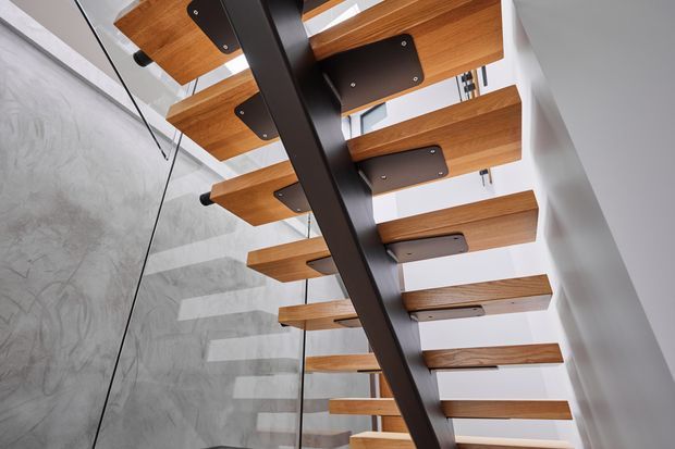 Close-up upward view of a modern open-riser staircase. Thick warm timber treads are cantilevered from a dark steel central spine, with visible dark metal mounting plates and a transparent glass balustrade beside a textured pale wall. Strong linear repetition and contrasting timber, black metal, glass, and concrete-like finishes create a contemporary architectural detail.