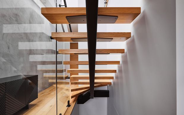 Interior architectural photograph of a contemporary floating staircase. Thick warm timber stair treads are cantilevered from a dark central structural spine, with slim black supports and a clear glass balustrade. A pale grey textured concrete or plaster wall and light timber flooring create a clean, refined composition.