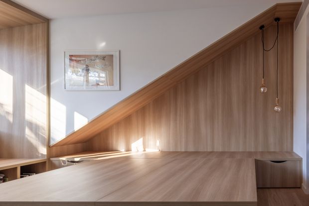 A warm, minimalist interior detail featuring a built-in timber study nook or desk beneath a dramatic sloping timber-lined wall and ceiling junction. Vertical timber panelling, a broad timber work surface, integrated shelving, and two hanging pendant bulbs create a calm, carefully crafted workspace. White walls and daylight from the left provide contrast, while a framed artwork adds a small decorative element.