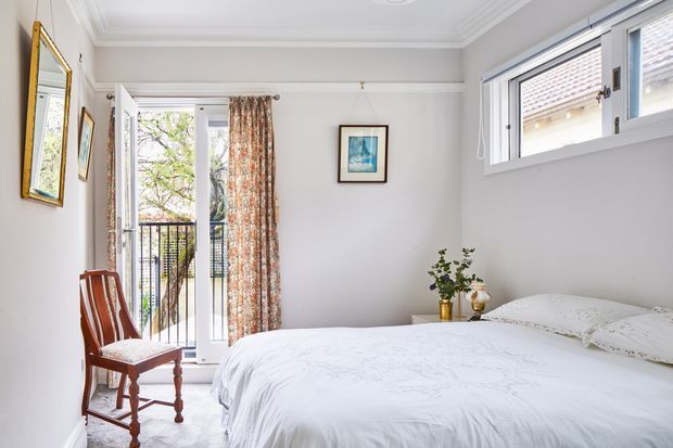 Bright traditional-style bedroom with a large white bed, pale walls, decorative ceiling cornice, framed artwork, a timber chair, and a glazed door opening onto a small balcony. Floral patterned curtains and classic trim add character, while the room has a calm, airy feel.