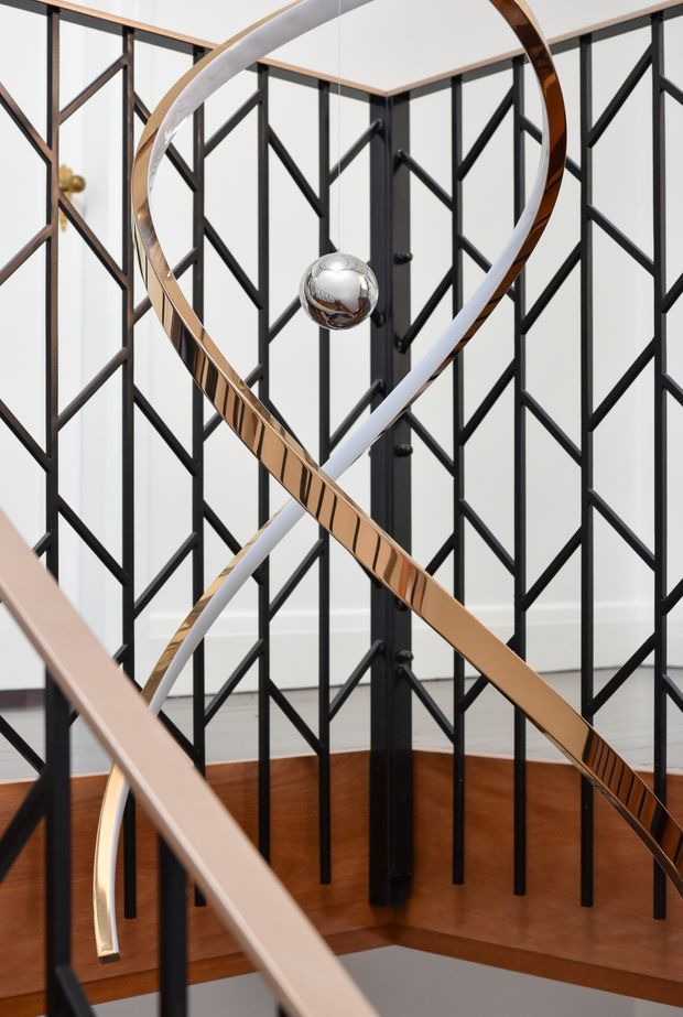 A sculptural stairwell interior featuring black geometric metal balustrade panels, warm timber wall and stair finishes, and an abstract suspended or integrated spiral artwork in white and polished bronze tones with a central chrome sphere. The space is refined and highly graphic, with strong vertical and diagonal lines and a visible stair void.