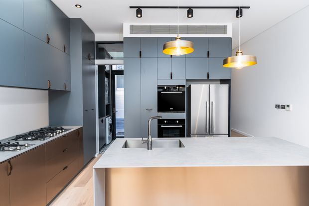 A contemporary kitchen with a large pale stone island and integrated sink, extensive blue-grey and bronze-toned cabinetry, a built-in oven, and a stainless steel double-door refrigerator. Two warm pendant lights hang above the island, complemented by black track lighting and a linear ceiling air vent. The room has a clean, refined fit-out with timber flooring and a bright neutral ceiling.