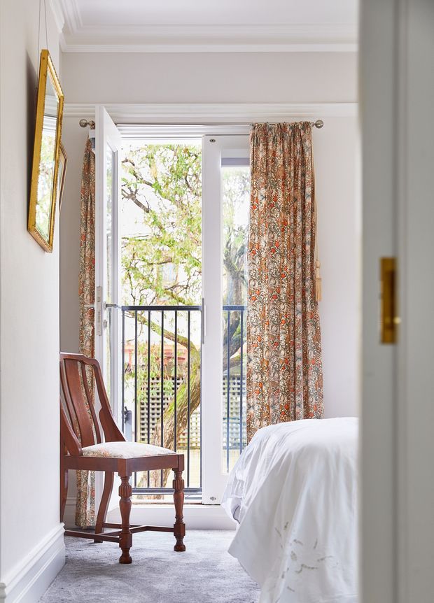 Bright, elegant bedroom viewed through a doorway, with a white upholstered bed partially visible in the foreground, a traditional dark timber chair, pale carpet, white decorative skirting and cornice, and tall French doors opening to a balcony. Patterned floral curtains in muted rust, green and beige frame the doors, while a gilt-framed mirror hangs on the wall.