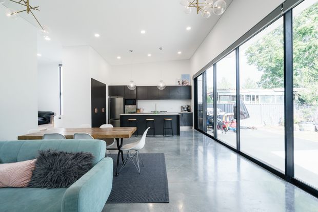 Bright open-plan contemporary living and kitchen area with polished concrete flooring, white walls and ceiling, dark built-in kitchen cabinetry, a timber dining table, pale blue sofa, and full-height black-framed glazing opening to an outdoor deck or patio. The room has a high ceiling, abundant natural light, and modern pendant and recessed lighting.