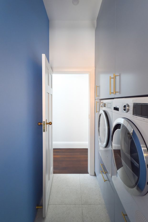 A narrow, bright laundry room with stacked front-loading white washer and dryer units along the right wall, integrated white cabinetry with brass-toned handles, pale tiled flooring, and a bold blue door and wall. An open white door leads to a hallway with dark timber flooring.