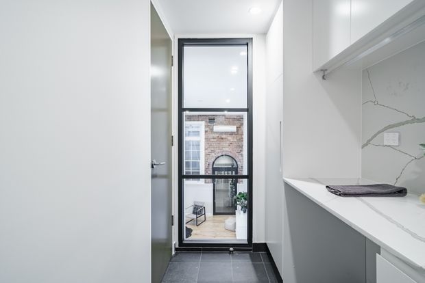 Bright, narrow contemporary utility or laundry space with white built-in cabinetry, a white stone-look benchtop and matching marble-veined splashback, open hanging rail, and dark tiled flooring. A full-height black-framed glazed door provides natural light and looks onto a brick courtyard or adjacent exterior area.