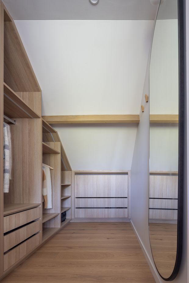 A compact, well-organised walk-in wardrobe with floor-to-ceiling light timber shelving, hanging rails, drawers and mirrored wardrobe doors. The room has a white ceiling and walls, timber-look cabinetry and warm timber flooring, with a clean contemporary fitted-out appearance.