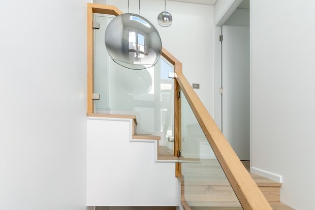 Bright contemporary stairway with pale timber stair treads and handrail, clear glass balustrade panels, and white walls and built-in stair structure. A spherical pendant light hangs above the stairs, with a hallway or room visible beyond the glazed stair enclosure.