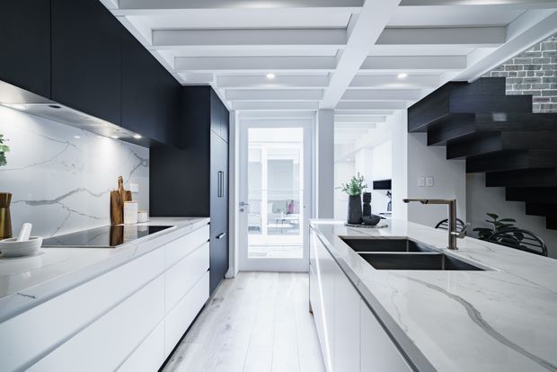 Bright contemporary kitchen with a long white island and integrated double sink, white veined stone benchtops and splashback, matte black cabinetry, and a dramatic open-riser black staircase. A white coffered or beam-lined ceiling, pale timber flooring, and a glazed rear door create a crisp, high-contrast interior.