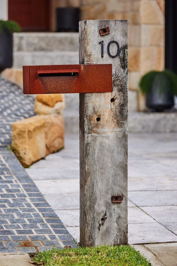 Close-up exterior detail of a freestanding weathered timber post supporting a rust-red metal letterbox and black house number 10. The post stands beside a paved entry path with contrasting patterned pavers, stone paving, rendered masonry, and landscaped planting visible around a residential entrance.
