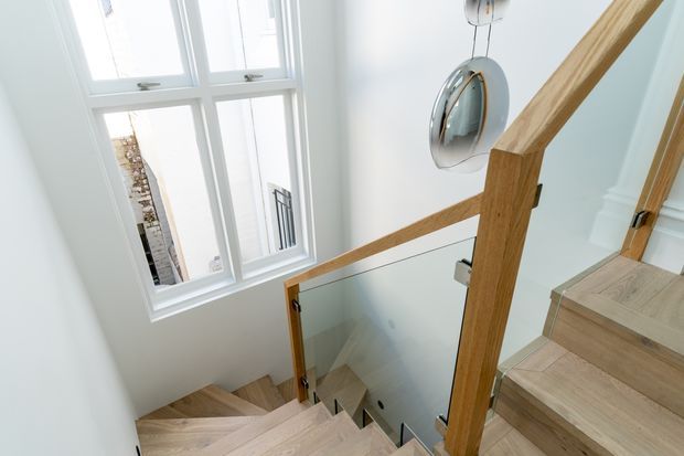 Bright contemporary stairway with pale timber treads and matching timber handrails, a clear glass balustrade, white walls and trim, and a tall multi-pane window bringing in abundant natural light. A modern pendant light hangs above the stairwell.
