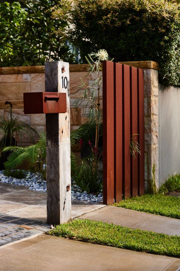 A landscaped residential front entrance viewed from the street, featuring a rough timber post with a rust-red metal letterbox and house number 10, beside a matching vertical rust-red metal gate. A low sandstone wall, concrete and stone-paver path, pebble garden bed, lush planting, and clipped hedges complete the entrance.
