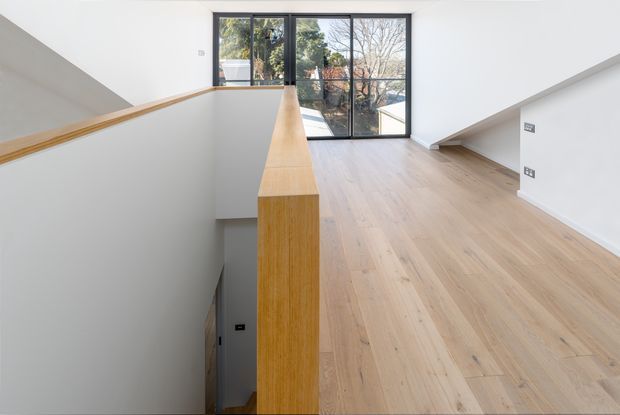 Bright minimalist upper-level landing or hallway overlooking a stairwell, with pale timber flooring and a matching timber-capped balustrade. White walls and angled soffits frame full-height black-framed glazing at the far end, looking toward trees and neighbouring buildings. The space is uncluttered and contemporary, with a strong sense of natural light and clean architectural lines.