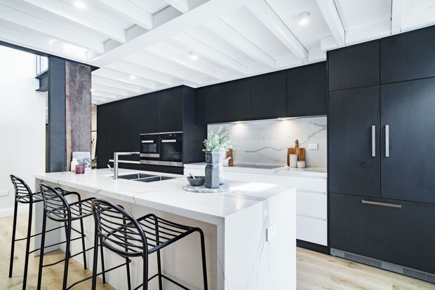 Bright contemporary kitchen featuring a large white stone island with integrated sink and seating for four, extensive matte black cabinetry, a white marble-look splashback and benchtop, integrated appliances, pale timber flooring, and a white exposed-beam ceiling. Black wire bar stools and understated accessories add contrast.