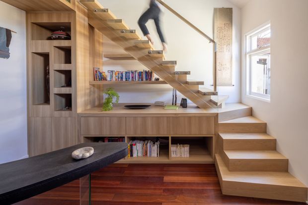 Bright contemporary interior featuring a custom timber library and storage wall integrated with an open-riser staircase. The stair has floating timber treads, a slim metal handrail, and clear glass balustrade, with built-in shelving, books, and a raised landing. Dark polished timber flooring contrasts with the lighter timber joinery and white walls. A partial dark island or table is visible in the foreground.