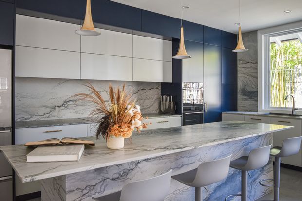 Contemporary kitchen with a large waterfall-edge stone island, integrated pale cabinetry, dark navy-blue cabinetry, a dramatic veined stone splashback, and four sculptural pendant lights. Grey bar stools line the island, while a window above the sink brings in natural light and greenery beyond.