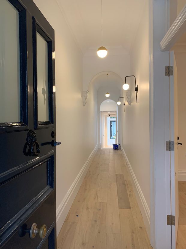 A long, narrow residential hallway viewed from an open dark-painted entrance door. The corridor has pale timber flooring, white walls and ceiling, decorative arched openings with ornate plaster brackets, classic white skirting and trim, and a series of simple pendant and wall-mounted lights. Open doorways lead to adjacent rooms, with daylight visible at the far end.