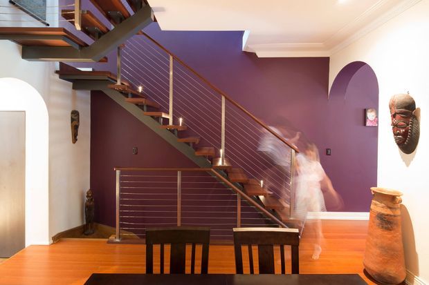 Interior view of a warm, contemporary stair hall and dining area. A floating timber staircase with open risers, dark metal stringers, and horizontal metal balustrade cables runs diagonally across a deep purple feature wall. Timber flooring, white walls with decorative ceiling cornices, arched openings, wall-mounted masks, and a dark dining table and chairs are visible. A blurred moving person appears near the stair landing.