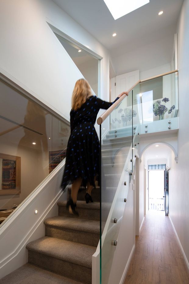 Bright, elegant interior view of a carpeted staircase and upper landing. The stair has a clear glass balustrade with a light timber handrail, white painted walls and trim, recessed ceiling lights, and a skylight. A hallway with pale timber flooring and an arched doorway is visible beyond, along with a framed artwork and decorative plants.