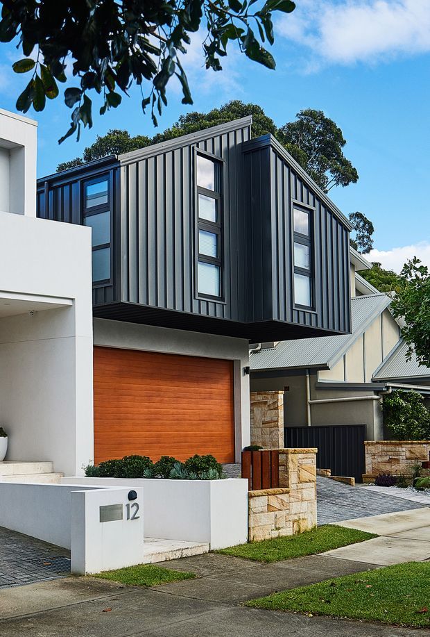 Street-facing view of a contemporary multi-storey home. The upper level projects over a garage and is clad in dark grey vertically ribbed metal panels, with tall narrow windows and dark frames. The ground level combines white rendered walls, a warm timber garage door, natural stone boundary walls, dark fencing, and a paved driveway. A neighbouring gabled dwelling and established trees are visible in the background.