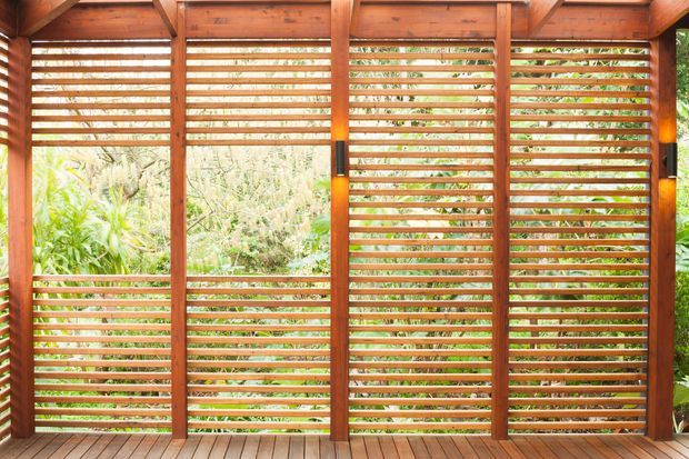 A timber-framed screened outdoor space with warm stained wood louvres and horizontal slatted panels enclosing three sides. The structure includes exposed beams, vertical posts, integrated warm wall lights, and a timber decking floor, with dense greenery visible beyond the screening.